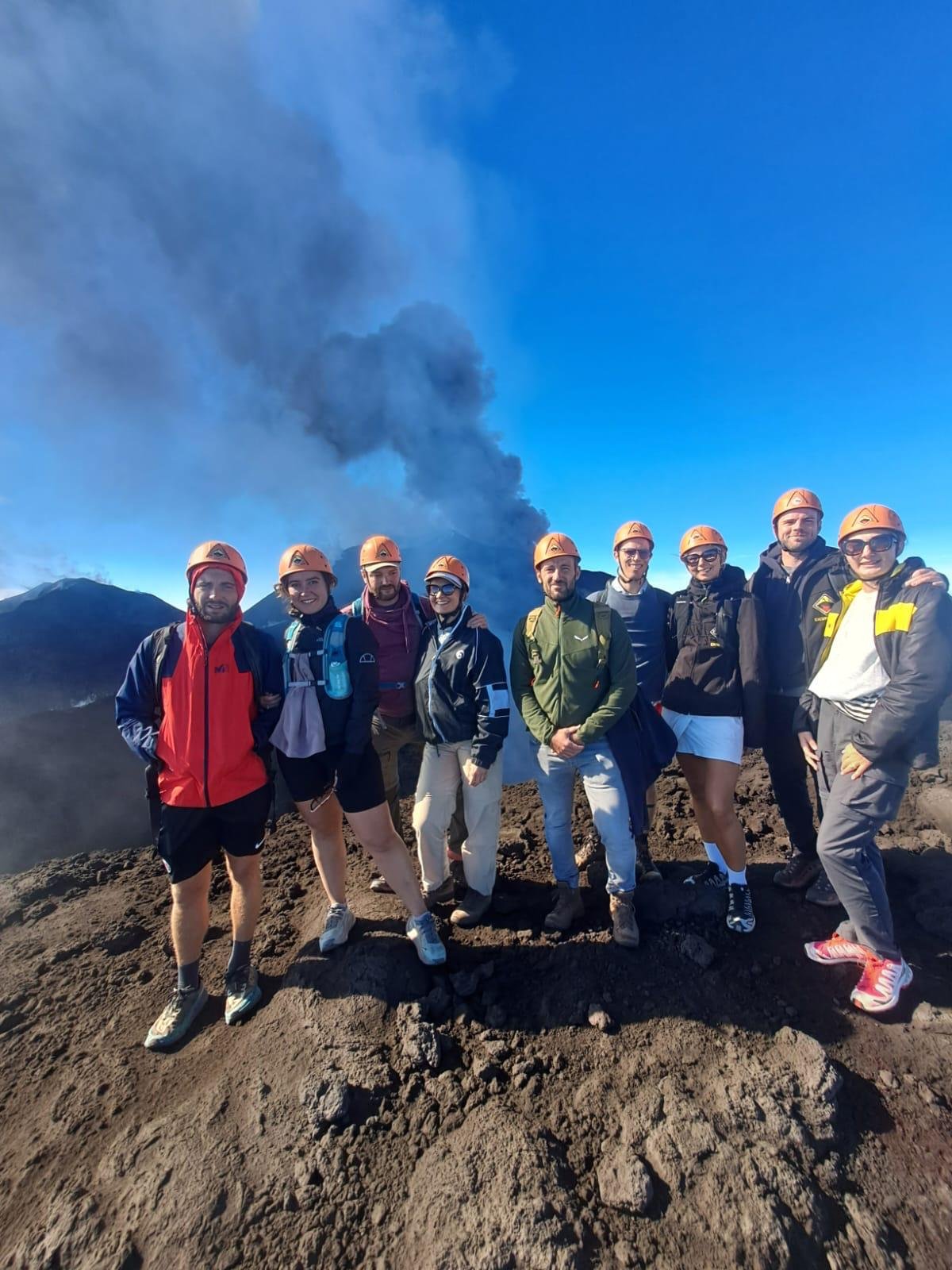 Groupe de randonneurs casqués en haute altitude sur l’Etna, avec le panache de cendres derrière eux
